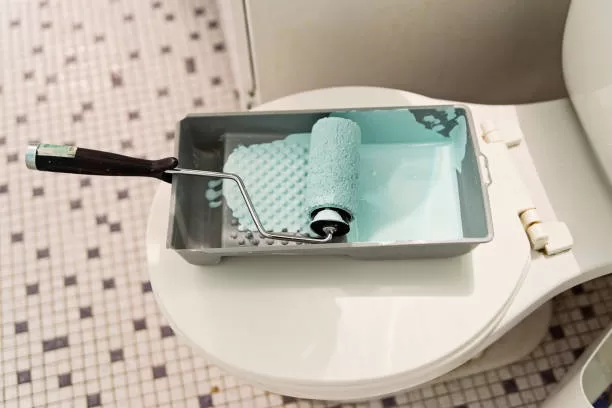 Step by step DIY in small appartement bathroom. A small pan of light blue paint is waiting on the toilet seat with a paint covered roll in it. Small tiles floor. Horizontal full length indoors shot with copy space.